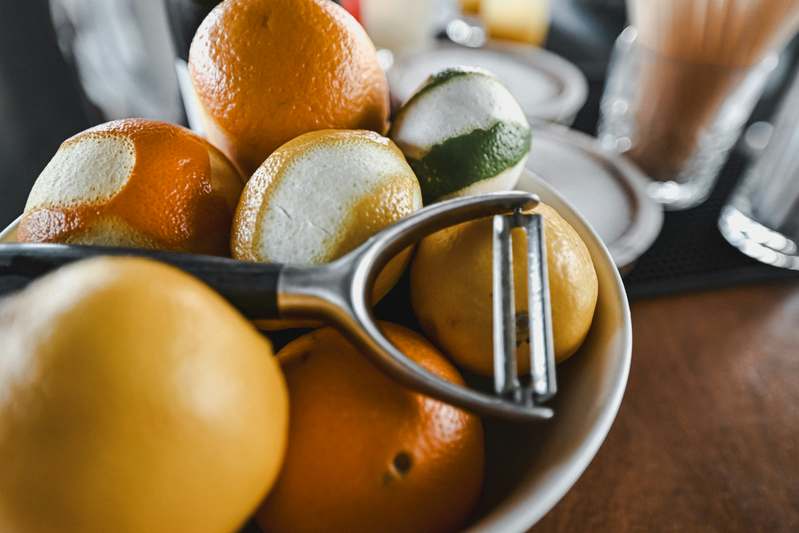 Side-by-side photo of five stainless steel and ergonomic potato peelers on a light wood countertop with peeled and unpeeled potatoes